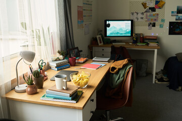 Modern workspace showing organized wooden desk with books, notebooks, coffee mug, bowl of chips, small plants, computer monitor on second desk, sunlight streaming through window