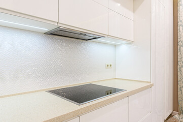 A modern, bright kitchen featuring white cabinets, a textured backsplash, and a speckled countertop. An electric stovetop and range hood are visible, and under-cabinet lighting adds a warm glow