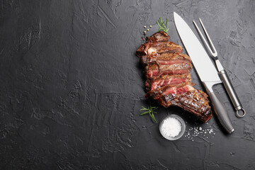 Tasty cut beef steak, salt, peppercorns, rosemary and cutlery on black table, flat lay. Space for...