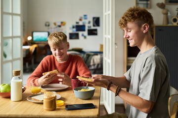 Two Caucasian teenage boys sitting at table eating breakfast together, smiling and holding toast, smartphone and breakfast foods visible on table, casual home setting © pressmaster