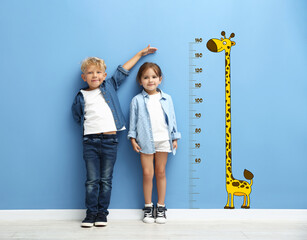 Cute little children measuring their heights near blue wall with scale and drawing of giraffe