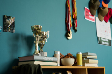 Books stacked on wooden shelf with trophies, medals hanging on blue wall, caps on hooks, snacks and drinks arranged nearby, suggesting teenager academic and athletic achievements