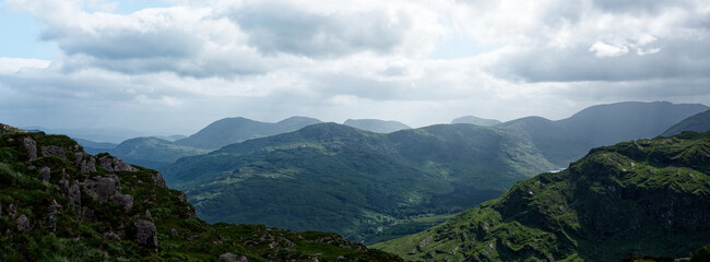 Fototapeta premium A panoramic view of rolling green hills and mountains, some capped with rocks, in the Gap of Dunloe in County Kerry, Ireland, under a cloudy sky during the daytime.