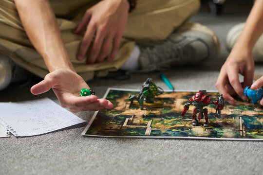 Two Caucasian teenage boys sitting on floor playing tabletop strategy game with dice and miniature robots, one holding green dice while other moving game piece on illustrated board