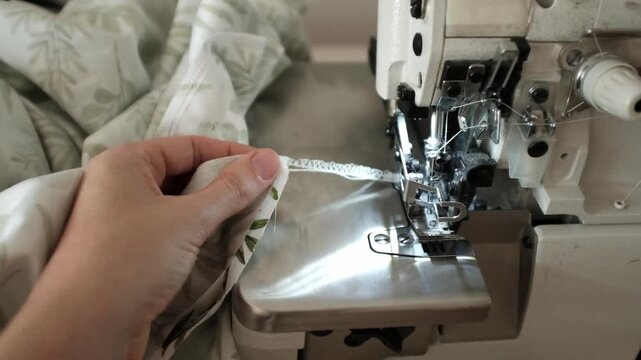 Woman sews bed linen or clothes on overlock sewing machine. Manufacture of textile goods. Professional sewing skills. Factory for production of clothing and textile goods.