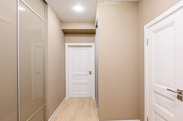 A neutral-toned hallway features light wood flooring, beige walls, and white paneled doors. Mirrored closet doors reflect the interior. A ceiling light provides illumination