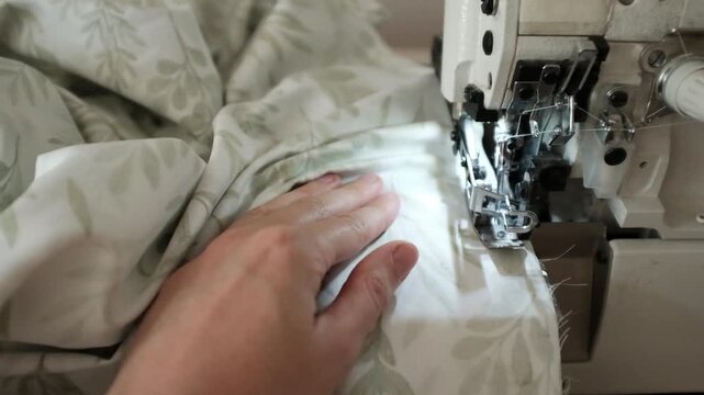Woman sews bed linen or clothes on overlock sewing machine. Manufacture of textile goods. Professional sewing skills. Factory for production of clothing and textile goods.