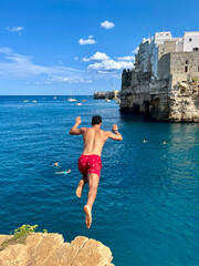 Mann springt von einer Klippe ins t&uuml;rkisfarbene Meer an der Adriak&uuml;ste, sonniger Sommertag mit klarem Himmel und wei&szlig;en H&auml;usern auf den Felsen