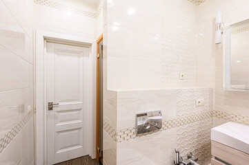 A bright bathroom with beige tiles, a white door, a built-in toilet, and modern fixtures. The design incorporates geometric patterns for added detail