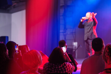 Audience watches vibrant stage performance as singer delivers show under red and blue lighting, crowd films on phones, energy high, intimate venue vibe
