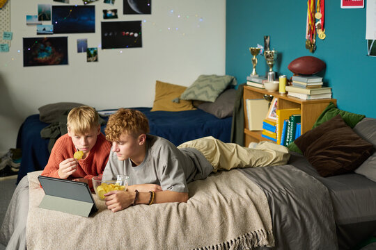 Two Caucasian teenage boys lying on bed watching tablet screen together, eating snacks from bowl, bedroom decorated with sports trophies and photos, brothers spending time together