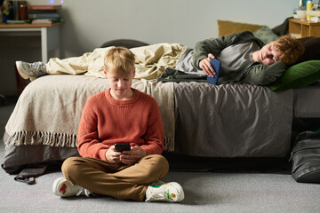 Caucasian teenage boy sitting on floor using smartphone while Caucasian teenage brother lying on bed holding phone, both engaging with devices in shared bedroom setting