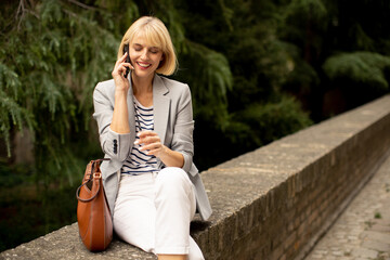 Woman talking on the phone while sitting on a stone wall surrounded by trees in a park setting