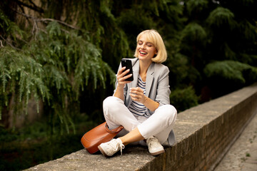 Woman sitting on a stone wall smiling at her phone in a park setting during daytime