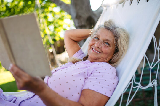 Older woman lying in a hammock and reading book, quiet time in garden. - Powered by Adobe