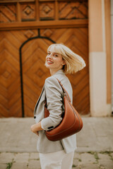 Young woman with short blonde hair smiles while walking down the street in a stylish blazer and carrying a brown handbag