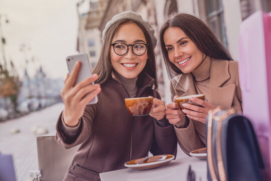 Young female friends women sitting outdoors in cafe drinking coffee and taking selfie photo vlogging blogging online on smartphone cellphone together, memory for social media