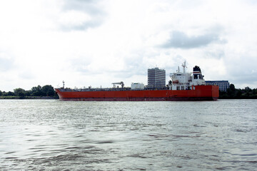 A large red cargo ship sails on a calm river with city buildings and cloudy sky in the background