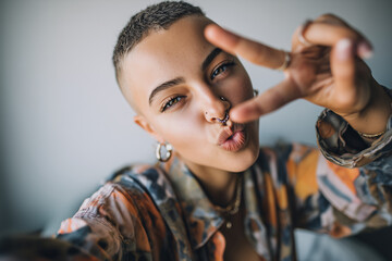 Playful confident person with buzzcut and septum piercing taking a close-up selfie, blowing a kiss and flashing a peace sign in colorful shirt