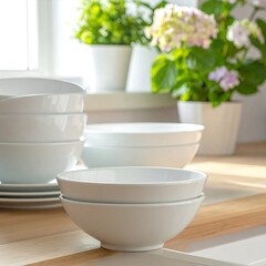 Stacked white bowls and plates on a light wooden kitchen counter,  with plants and sunlight