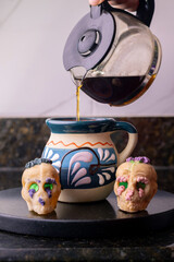 Hand pouring hot coffee into a traditional mexican cup beside two colorful sugar skulls. day of the dead celebration