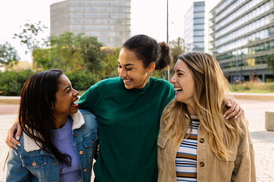 Three happy multiracial young women laughing outdoors. United female best friends having fun walking in city street. Community and international friendship concept