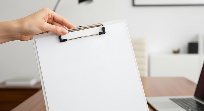 A hand is holding a blank clipboard in front of a desk with a laptop, ready for notes or a document to be added, creating a workspace scene