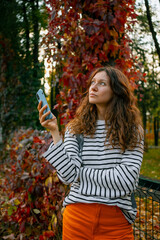 Woman holding smartphone in hands, looking up thoughtfully at vibrant autumn leaves, connecting with nature and technology in a beautiful fall park setting, enjoying mindful moments outdoors