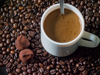 A perfect coffee break scene. A white cup of fresh coffee with a spoon is surrounded by roasted coffee beans, with two delicious, cocoa-dusted chocolate truffles