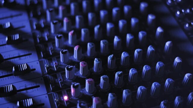 Close-up pan across an audio mixing console with knobs, faders, and buttons under cool blue lighting. The shot transitions from sharp detail to a soft focus bokeh effect, showing professional gear.