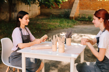 2 Girls sculpt clay in an outdoor workshop. Clay art workshop outdoors. Two women enjoy a pottery class outdoors, shaping clay with smiles and creativity.