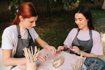 2 Girls sculpt clay in an outdoor workshop. Clay art workshop outdoors. Two women enjoy a pottery class outdoors, shaping clay with smiles and creativity.