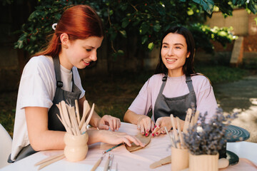 2 Girls sculpt clay in an outdoor workshop. Clay art workshop outdoors. Two women enjoy a pottery class outdoors, shaping clay with smiles and creativity.