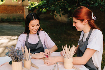 Two women enjoy a pottery class outdoors, shaping clay with smiles and creativity. 2 Girls sculpt clay in an outdoor workshop. Clay art workshop outdoors.