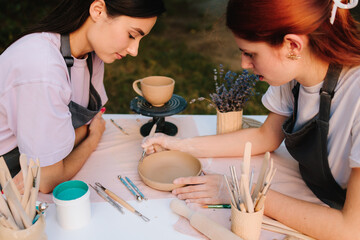 Outdoor pottery class. Two women enjoy outdoor pottery class, sitting at table with clay and carving tools while learning ceramic crafting. Outdoor pottery session shows students working with clay.