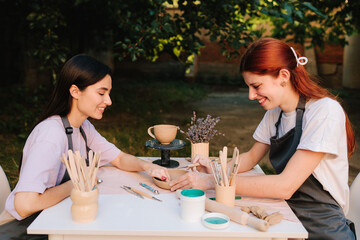Outdoor pottery class. Two women enjoy outdoor pottery class, sitting at table with clay and carving tools while learning ceramic crafting. Outdoor pottery session shows students working with clay.