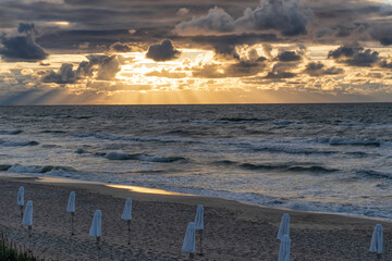 A dramatic sunset over the sea. Rays of sun break through dark clouds. Umbrellas are rolled up on the beach. Background. Landscape.