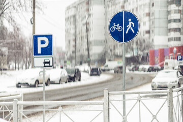 Two blue traffic signs, one for parking and one for shared pedestrian and bicycle path, stand in a blurred, snow-covered city street with cars and buildings.