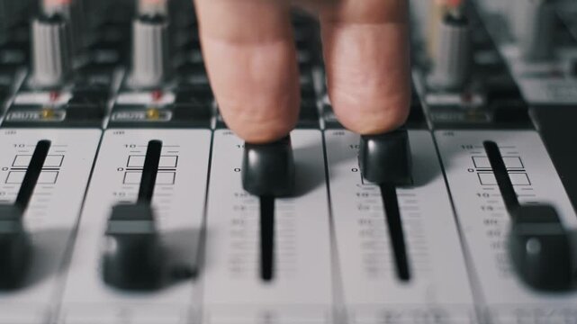 Close-up side angle of fingers pushing faders up on professional audio mixing console, adjusting volume levels in recording studio. Shallow depth of field highlights black fader caps and numbered