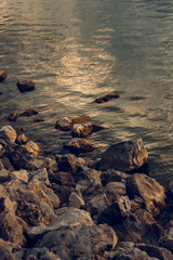 A close-up view of large, dark rocks piled along a shore next to tranquil water reflecting warm, soft sunlight during sunset or sunrise.