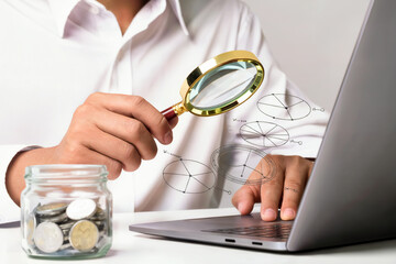 Person analyzing financial data on laptop with magnifying glass and jar of coins nearby on desk