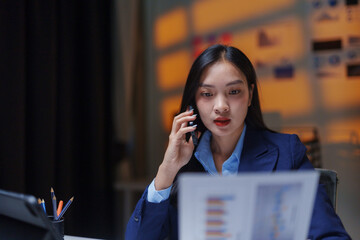 Young Asian businesswoman using mobile phone and reviewing financial documents in a dimly lit office, working late hours