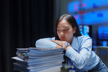 Young Asian businesswoman feeling tired and stressed, resting head on a tall stack of documents late at night in an office