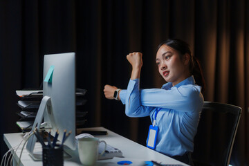 Asian businesswoman stretching her arms while working late at her desk in a dark office, needing a...