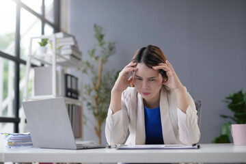 Young asian businesswoman feeling overwhelmed and stressed from workload, experiencing a headache while working at desk