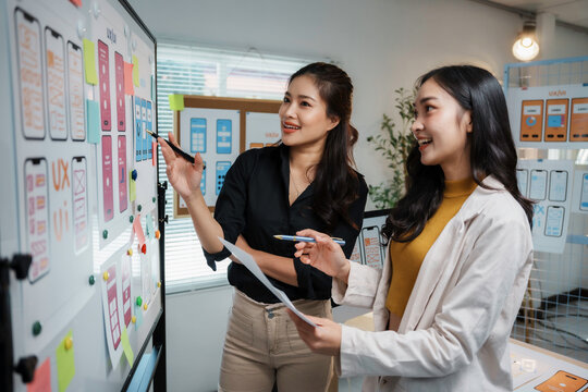 Two Asian women UXUI designers collaborating on a whiteboard, brainstorming and planning mobile app user interface wireframes and sticky notes