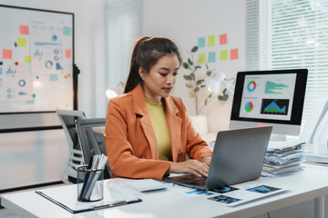Young Asian businesswoman working on laptop, analyzing financial data and charts on computer screens and documents, managing corporate strategy