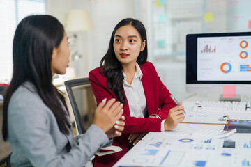 Two Asian businesswomen collaborating on a project, analyzing charts and graphs on documents and a computer screen in an office