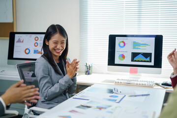 Asian businesswoman clapping hands, smiling with colleagues at a business meeting, celebrating achievement with data charts on screens