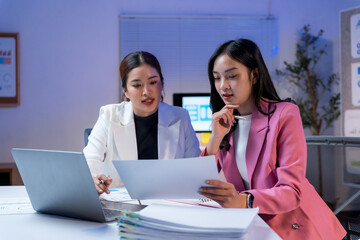 Two Asian businesswomen collaborating on a project, analyzing documents and using a laptop in a modern office at night